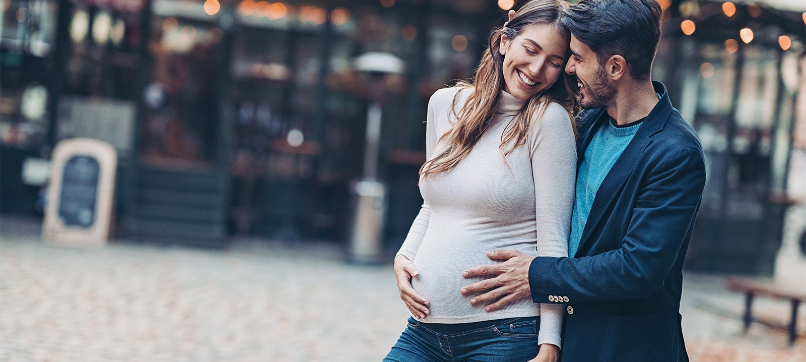 A young couple smiles as the man and woman place a hand on the woman's pregnant stomach in a city courtyard setting